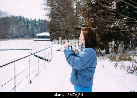 femme en hiver avec le téléphone prend des photos de la nature dans le parc par temps froid Banque D'Images