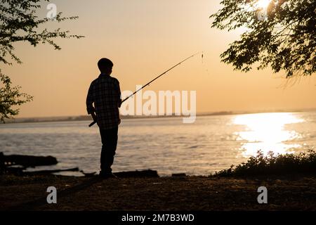 Silhouette d'un garçon pêchant sur un lac en été au coucher du soleil Banque D'Images