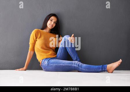 Dire oui à la détente. Une jeune femme attirante s'est penchée contre un mur gris tout en étant assise sur le sol. Banque D'Images