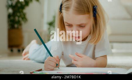 Mignon concentré caucasien petite fille dessin couleur crayon allongé sur le sol à la maison jouer seul. Talent préscolaire enfant fille enfant enfant bébé colorant Banque D'Images