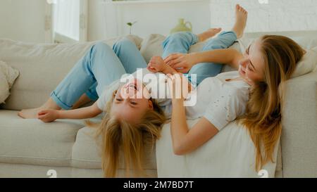 Mère de race blanche mère avec fille couché sur canapé canapé dans le salon momie tickle gamin fille riant chatouillement enfant bébé sincère rire drôle profiter Banque D'Images