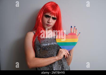 Portrait d'un homme transgenre tenant un coeur de drapeau arc-en-ciel sur fond gris. Banque D'Images