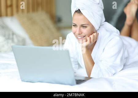 Bonne adolescente couchée sur un lit vérifiant ordinateur portable après avoir pris la douche à la maison Banque D'Images