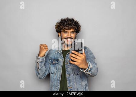 Heureux indien homme investisseur gagnant en utilisant le téléphone mobile isolé sur fond gris. Un homme ethnique enthousiaste tenant un smartphone se sent surpris par le bett Banque D'Images