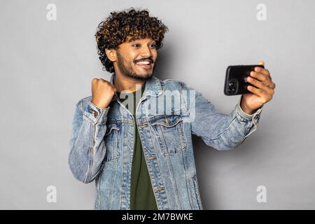Heureux indien homme investisseur gagnant en utilisant le téléphone mobile isolé sur fond gris. Un homme ethnique enthousiaste tenant un smartphone se sent surpris par le bett Banque D'Images