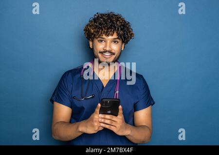 Portrait d'un joli médecin indien avec stéthoscope tenant son téléphone tout en restant isolé sur fond bleu. Banque D'Images