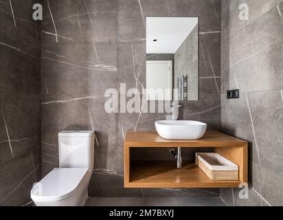 Dressing avec murs en marbre gris foncé avec touches blanches, petit lavabo ovale en céramique sur meuble en bois et miroir sur mur qui se reflète Banque D'Images