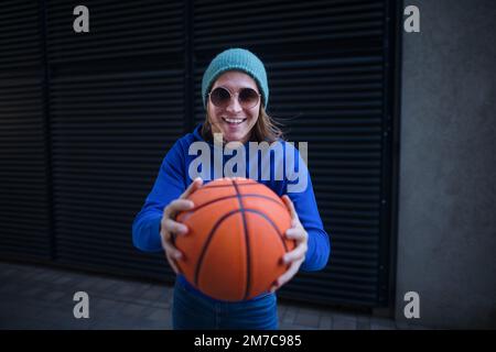 Portrait d'un jeune homme avec chapeau et lunettes de soleil tenant le ballon de basket-ball, extérieur en ville. Culture de la jeunesse. Banque D'Images