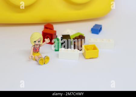 Une fille aux cheveux blonds agite. Elle est assise à côté de blocs LEGO dispersés ann un bloc LEGO géant en arrière-plan. Banque D'Images