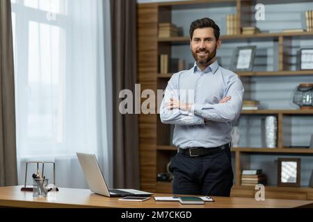 Portrait d'un homme d'affaires au bureau, d'un homme en chemise souriant et regardant l'appareil photo, d'un patron mûr avec une barbe aux mains déchiqueuses debout sur le lieu de travail à l'intérieur du bâtiment. Banque D'Images