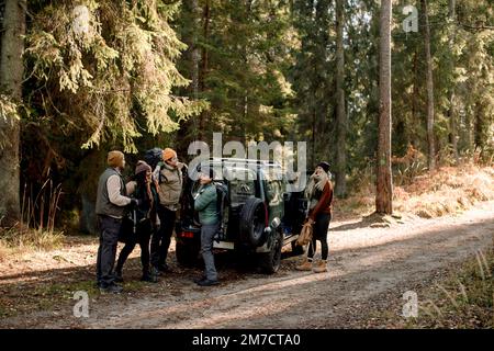 Des amis multiraciaux, hommes et femmes, se parlent les uns avec les autres tout en se tenant près d'un véhicule tout-terrain sur une route de terre contre l'arbre Banque D'Images