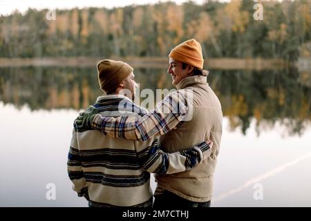 Des amis masculins parlent les uns avec les autres tout en se tenant avec le bras autour contre le lac Banque D'Images