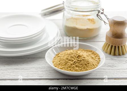 Concept de lavage de la vaisselle avec de la moutarde en poudre. Nettoyez les assiettes blanches avec un bol et un bol de poudre de moutarde jaune et une brosse à vaisselle en bois. Banque D'Images