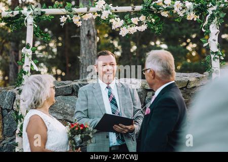 Couple senior échangeant des voeux tout en regardant le ministre debout contre le mur avec la décoration de fleur pendant le mariage céré Banque D'Images