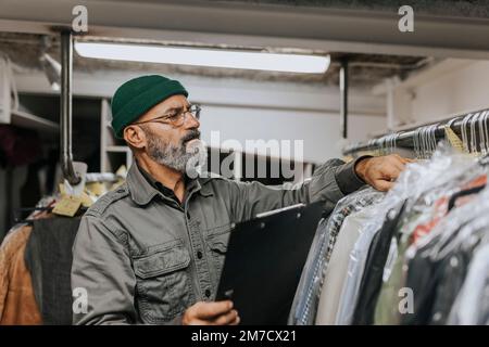 Tailleur mâle barbu avec presse-papiers pour vérifier les vêtements suspendus sur le rack dans l'atelier Banque D'Images