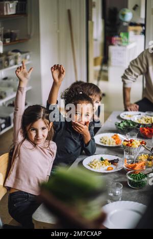 Filles et garçons heureux avec les mains levées à la table de salle à manger pendant le petit déjeuner à la maternelle Banque D'Images