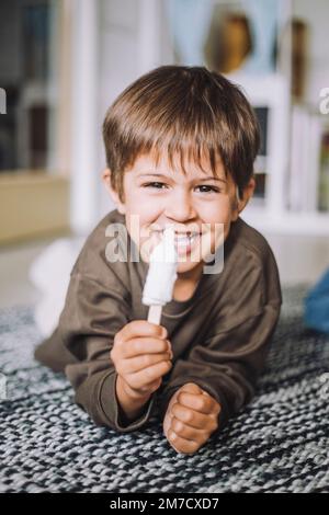 Portrait d'un garçon souriant ayant de la glace tout en étant allongé sur un tapis Banque D'Images