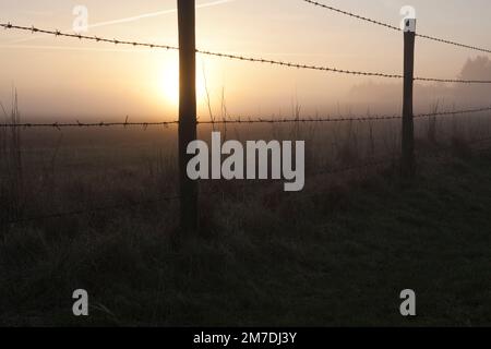Les couchers de soleil sur un champ dans le pays de jeter dans une silhouette de barbelés qui traverse les champs. Banque D'Images
