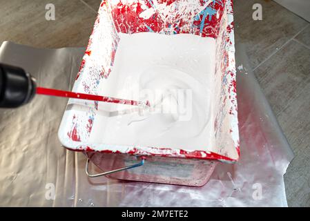 Un seau en plastique ouvert avec peinture blanche et un agitateur debout sur le sol. Banque D'Images