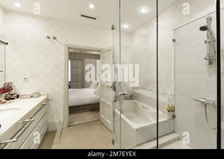 Salle de bains avec mur recouvert de mosaïque blanche, lavabo en marbre blanc à deux vasques avec tiroirs, baignoire et cabine de douche séparées avec porte en verre et p Banque D'Images