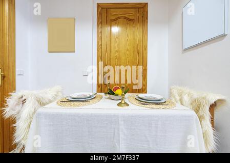 Table à manger pour deux avec nappe et chaises en laine polaire blanche à côté d'une porte en pin verni Banque D'Images