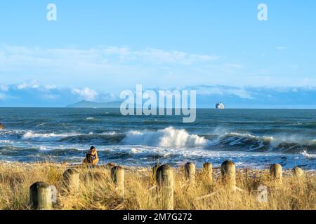 Mont Maunganui Nouvelle-Zélande - 9 janvier 2022: Femme sur la plage comme bateau de croisière arrivant au Mont Maunganui à l'horizon au-delà des vagues de rupture Banque D'Images