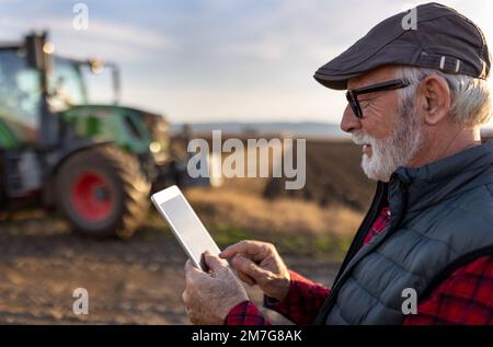 Un agriculteur mûr enthousiaste travaillant sur une tablette devant le tracteur dans les champs Banque D'Images