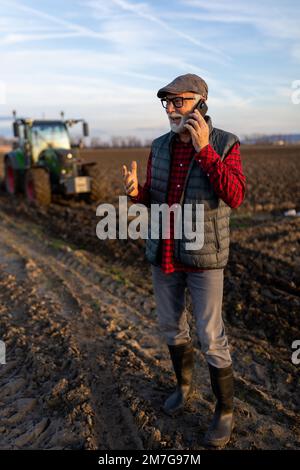 Un agriculteur mûr enthousiaste qui parle sur un téléphone portable devant le tracteur dans les champs Banque D'Images
