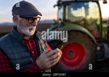Ferme mûre souriante regardant le téléphone mobile devant le tracteur dans le champ. Concept d'agriculture intelligente Banque D'Images