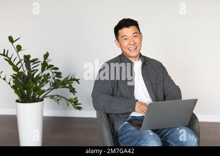 Souriant beau homme asiatique mûr assis dans un fauteuil dactylographiant sur l'ordinateur, regardant l'appareil photo, appréciez le travail Banque D'Images