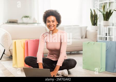 Shopaholic, blogueur de mode. Jeune femme souriante de race mixte dactylographiant sur un ordinateur portable au sol dans le salon Banque D'Images