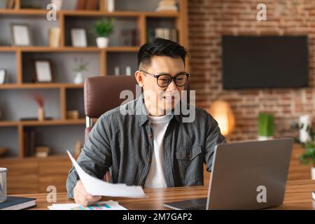 Souriant beau adulte asiatique homme en lunettes travaille avec des documents sur le lieu de travail, a la réunion dans l'intérieur de bureau Banque D'Images