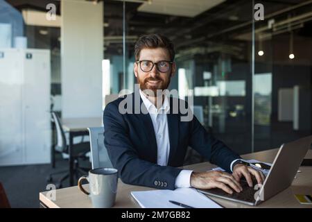 Portrait d'un homme d'affaires d'âge moyen confiant assis sur une table de travail au bureau, en tapant sur le clavier, en regardant l'appareil photo Banque D'Images