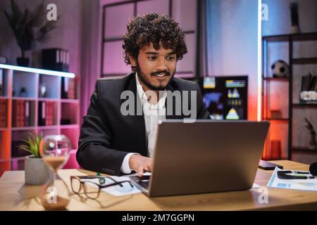 Beau expérimenté élégant et élégant gérant, courtier, homme d'affaires, portant un costume assis à table, utilisant un ordinateur portable le soir dans un bureau moderne. Banque D'Images