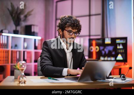 Homme à barbe confiant en costume et lunettes habillée assis au bureau avec ordinateur portable moderne et regardant l'écran avec une expression faciale sérieuse. Concept de personnel et de travail de bureau. Banque D'Images