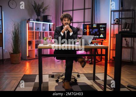 Portrait complet d'un homme barbu souriant en costume habillé assis au bureau avec un ordinateur portable moderne et regardant l'appareil photo le soir au bureau. Concept de personnes, échéance et travail de bureau. Banque D'Images