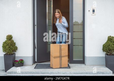 Jeune femme excitée ouvrant la porte d'entrée et voyant le paquet sur le tapis de porte Banque D'Images