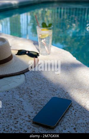 Verre rafraîchissant d'eau, chapeau de paille, lunettes de soleil et téléphone à la piscine Banque D'Images
