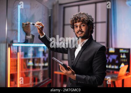 Maîtrise professionnelle compétente en gestion de projets dans un bureau moderne. Gros plan d'un homme d'affaires concentré dans des notes d'écriture de vêtements formels, des tableaux et des tâches de travail sur le mur de verre. Banque D'Images