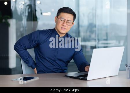 Un jeune étudiant asiatique est assis dans le bureau à un bureau avec un ordinateur portable. Il ressent une douleur sévère dans le dos, grimaces de la douleur et de la fatigue. Banque D'Images