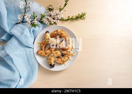 Biscuits en croissant d'amande sur une assiette, branche de fleur de ressort et serviette bleue sur une table en bois clair, grand espace de copie, vue d'en haut Banque D'Images