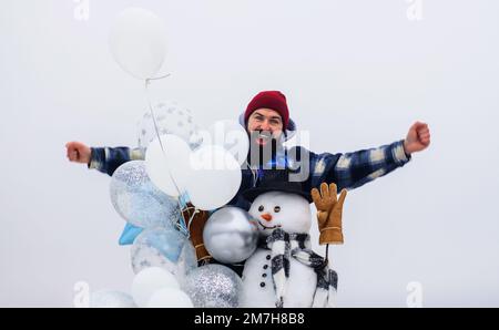 Joyeuses vacances. Homme barbu en hiver avec bonhomme de neige en chapeau, écharpe et moufles avec ballons d'air. Banque D'Images