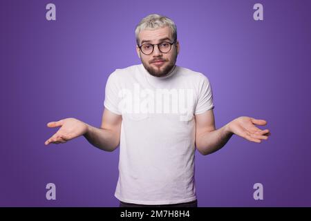 Portrait de beau barbu jeune homme debout avec les bras relevés, ne sais pas quoi faire, isolé sur fond violet. Je ne sais pas. Banque D'Images