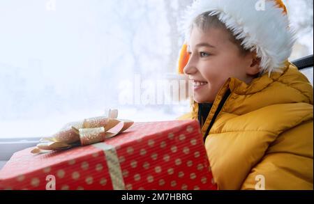 Vue latérale d'un petit enfant portant un chapeau de Noël, voyageant en Tarnsport public, assis dans le bus, souriant. Adorable enfant tenant une boîte cadeau, regardant vers l'avenir. Concept de vacances d'hiver. Banque D'Images