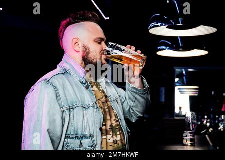 Un jeune homme charmant boit de la bière au bar. Un punk d'apparence informelle tient un verre de bière dans la main contre le fond d'un bar de nuit. Banque D'Images