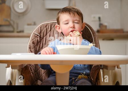 Un enfant drôle mange une pomme râpée avec sa bouche pleine tout en étant assis sur une chaise de cuisine. Bébé garçon affamé, il a de la nourriture dans sa bouche, de l'humour. Enfant âgé Banque D'Images