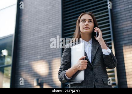 Belle femme caucasienne parlant sur un téléphone mobile tout en allant au bureau avec un ordinateur portable. Jeune fille indépendante appelle aux clients Banque D'Images