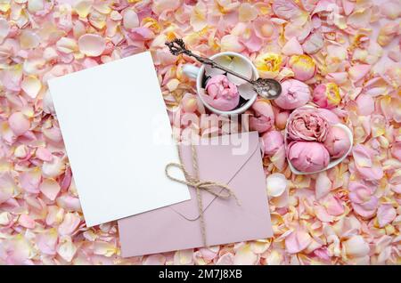 Fond de rose rose bourgeons et pétales avec enveloppe et carte vierge au centre et une tasse de thé avec rose et cuillère à thé. Pose à plat, vue de dessus. Papillon Banque D'Images