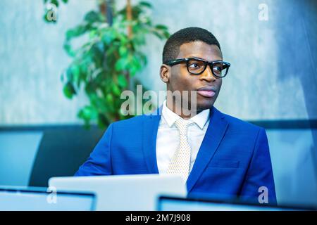 Homme d'affaires afro-américain en costume bleu et lunettes stylées qui regardent loin tout en utilisant un ordinateur portable pendant le travail dans un bureau moderne Banque D'Images