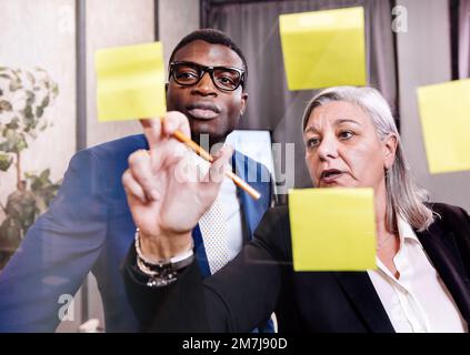 Femme d'affaires mûre pointant vers des notes adhésives et expliquant des données à des collègues noirs tout en se tenant derrière une fenêtre dans un bureau moderne Banque D'Images